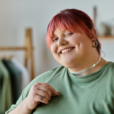 smiling woman with pink hair wearing a green shirt looking at the camera in a cozy indoor setting with fashion items related to styles for four