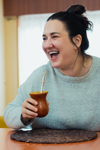 woman joyfully laughing while enjoying traditional drink sitting at a table with mate and cozy sweater