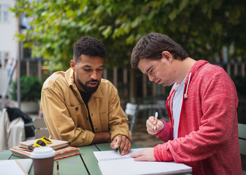 two young men studying together outdoors engaged in discussion focused on a notebook and textbooks with a coffee cup on the table includes concepts related to 14 educational topics